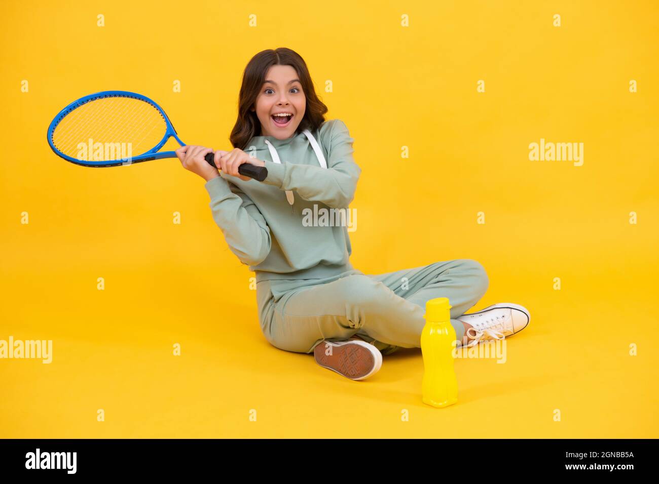 bambino felice sedersi con acqua bottiglia swing racket tennis per colpire palla su sfondo giallo, attività. Foto Stock