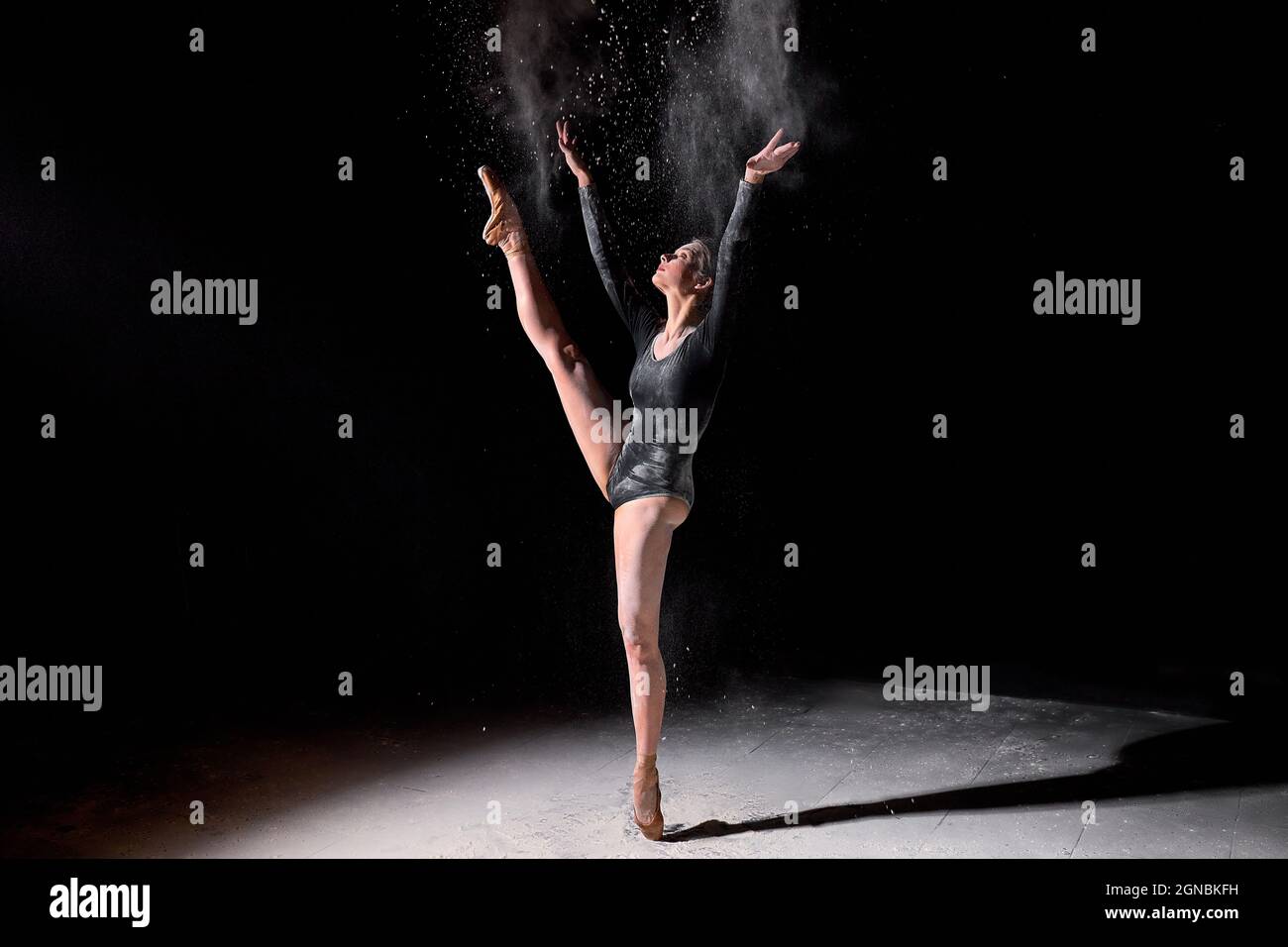 Ballerina flessibile donna danzante e spolverare la farina su sfondo nero studio, indossando tuta nera e scarpe a punta. Ritratto completo di attra Foto Stock