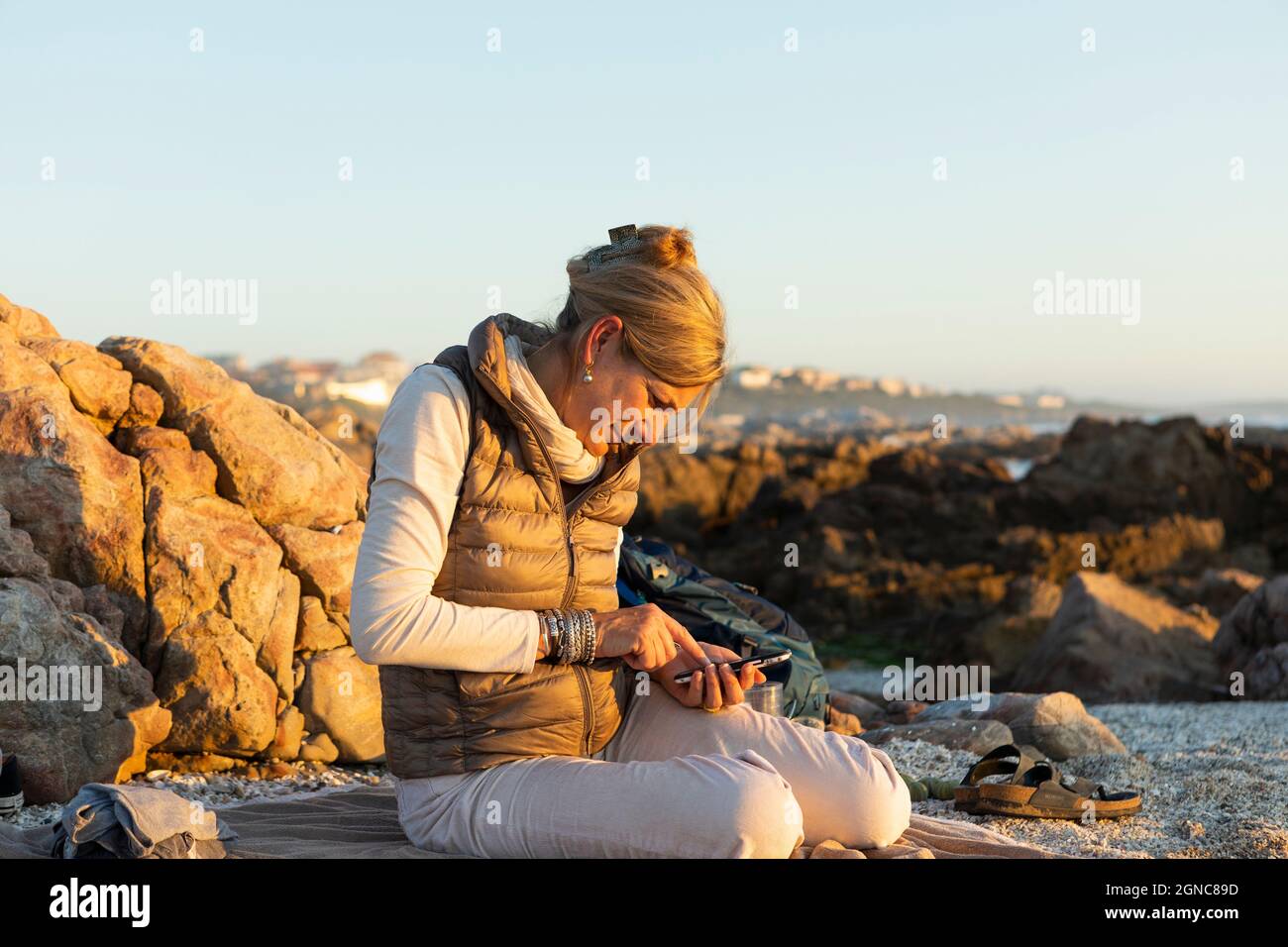 Una donna adulta seduta sulla spiaggia usando il suo smartphone al De Kelders al tramonto. Foto Stock