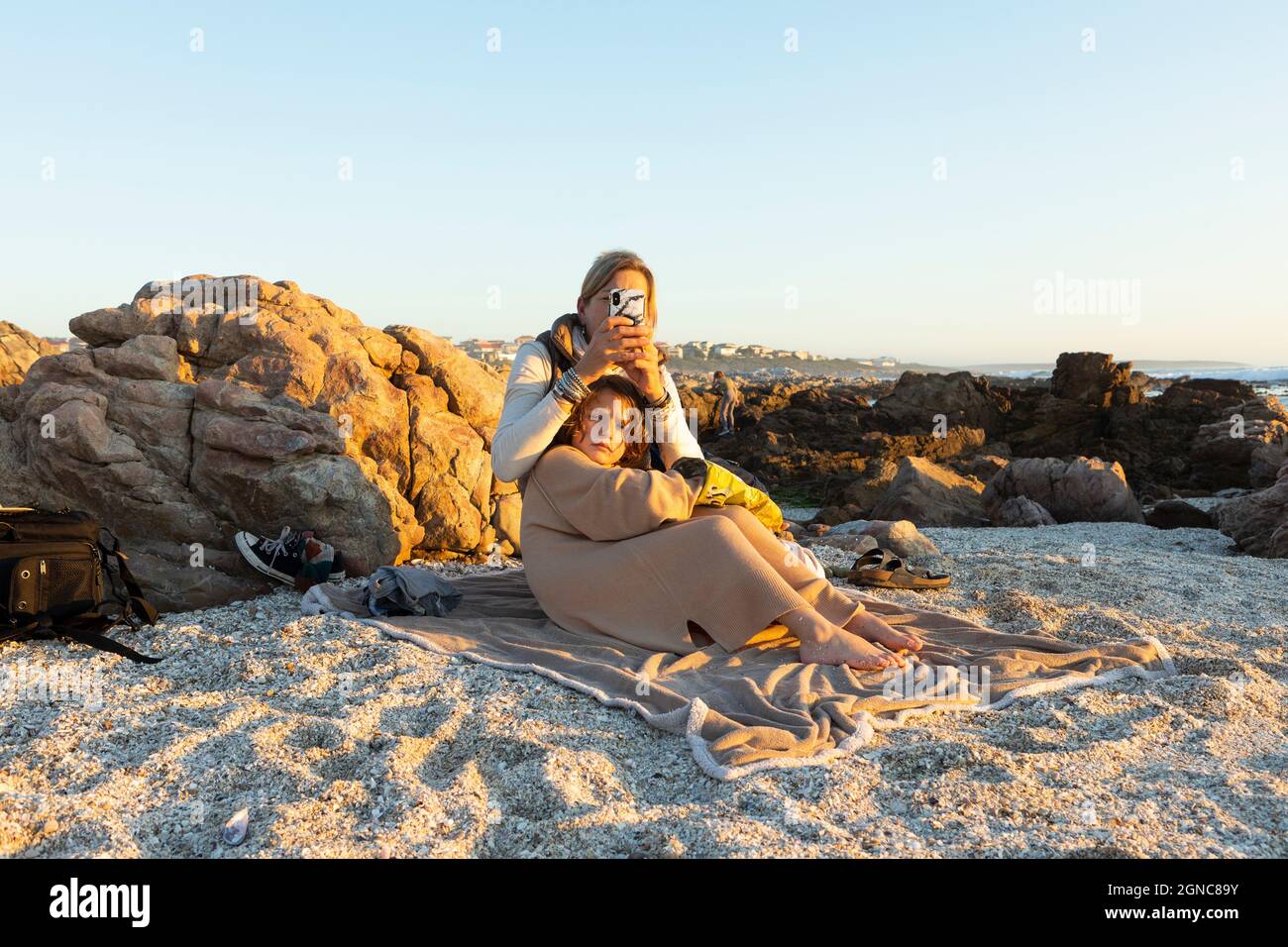 Una donna e suo figlio seduti sulla sabbia tra le rocce sulla riva di De Kelders al tramonto. Foto Stock