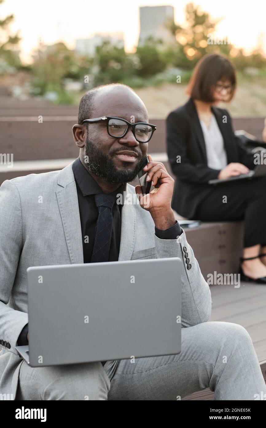 Ritratto verticale medio di bell'uomo afroamericano portatore di abito elegante seduta su panca all'aperto con computer portatile che parla al telefono con col Foto Stock