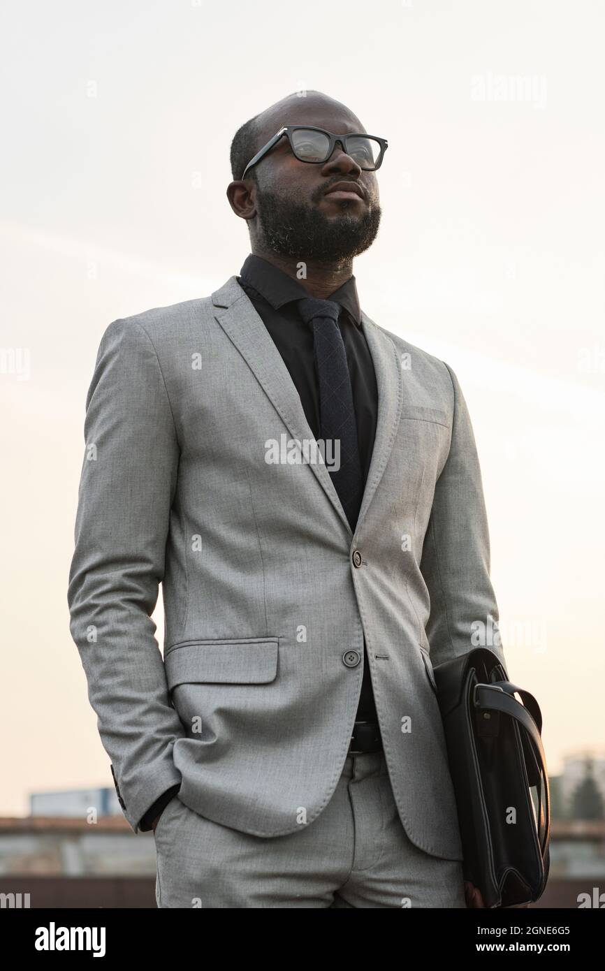 Inquadratura verticale ad angolo basso di un uomo afro-americano che indossa un elegante abito grigio e occhiali in piedi all'aperto guardando lontano Foto Stock