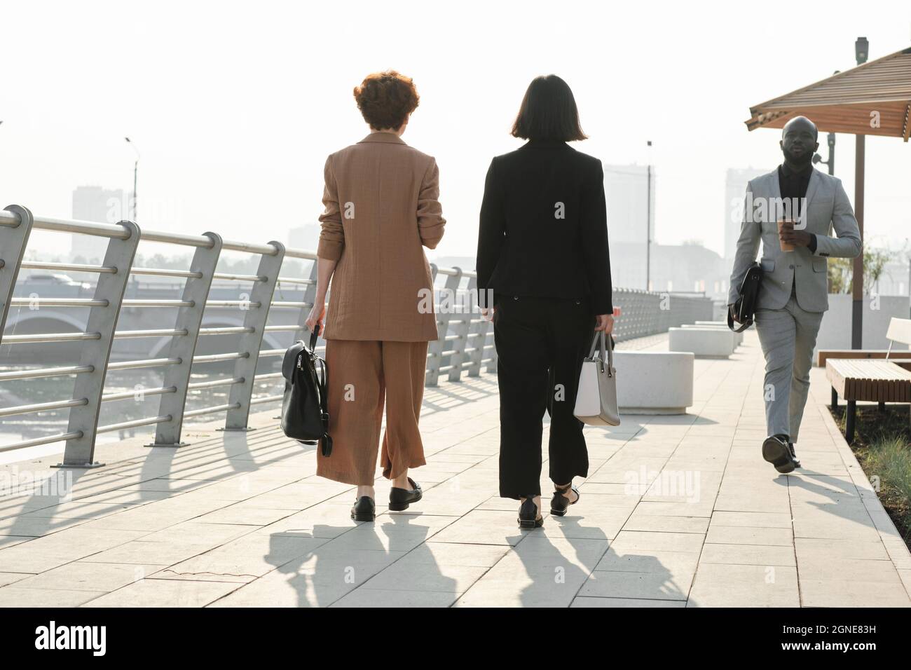Ampio scatto orizzontale di donne irriconoscibili e uomo afroamericano indossando eleganti abiti formali camminando lungo la riva del fiume nella città moderna Foto Stock