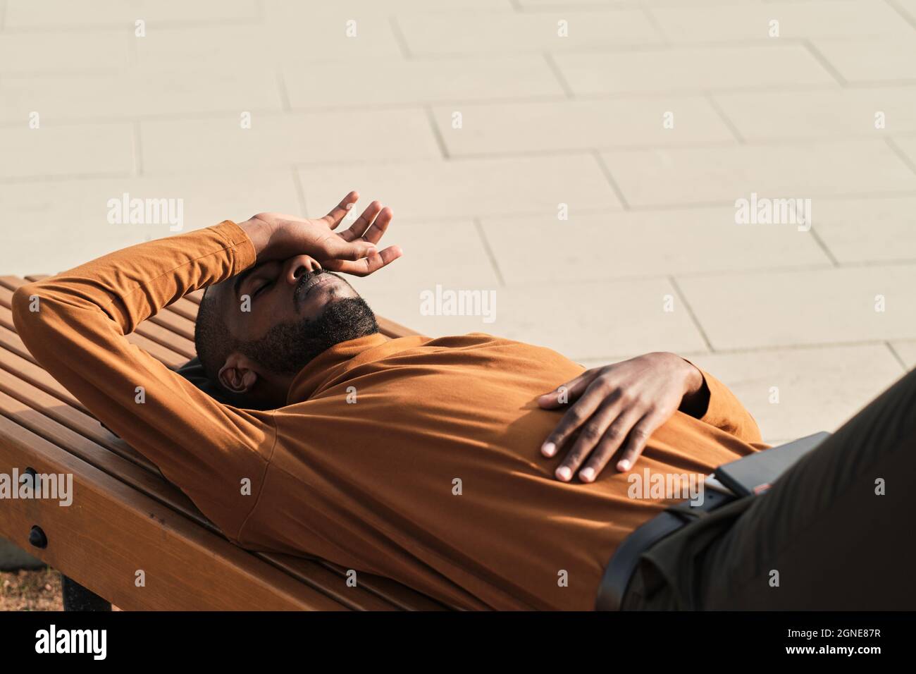Orizzontale alto angolo di ripresa del giovane afro americano stanco rilassarsi su panca di legno all'aperto nella soleggiata giornata estiva Foto Stock