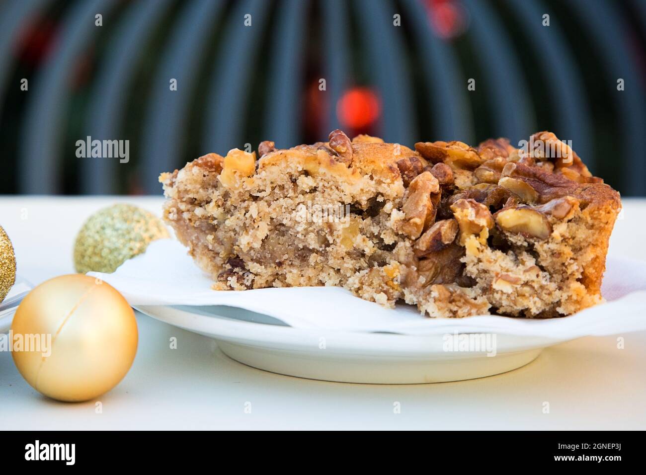 Palla di Natale d'oro e un pezzo di torta al caramello con noci. Tavolo bianco, sedia grigia e albero di nuovo anno con giocattoli rossi in background fuori fuoco Foto Stock