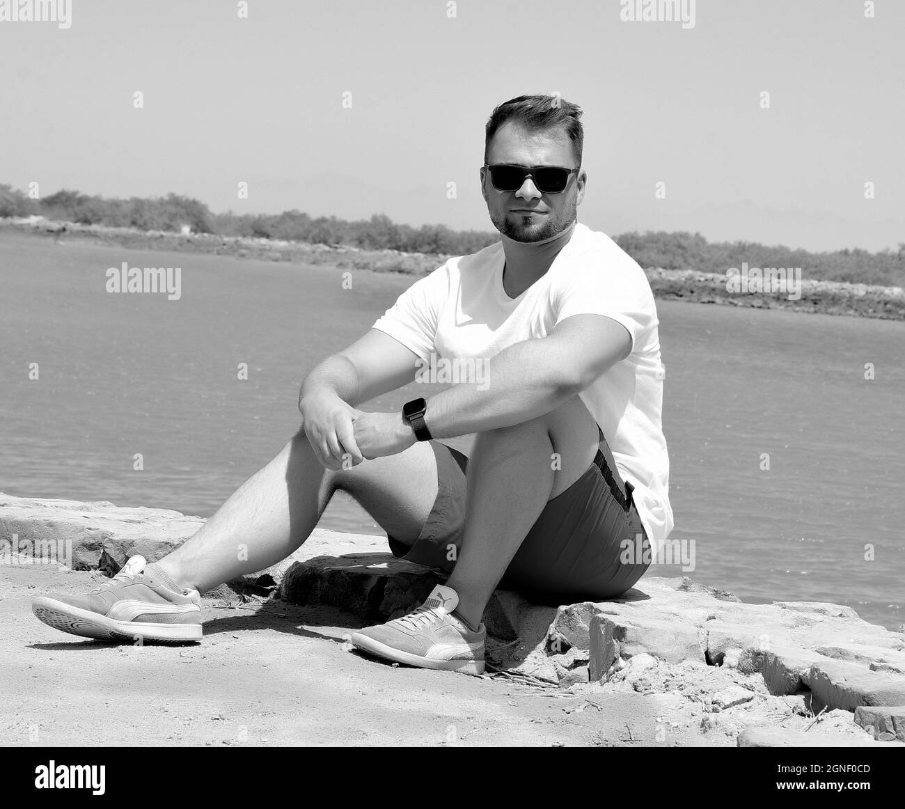 il giovane uomo si siede di fronte al mare in vacanza con abiti corti e indossa occhiali da sole Foto Stock