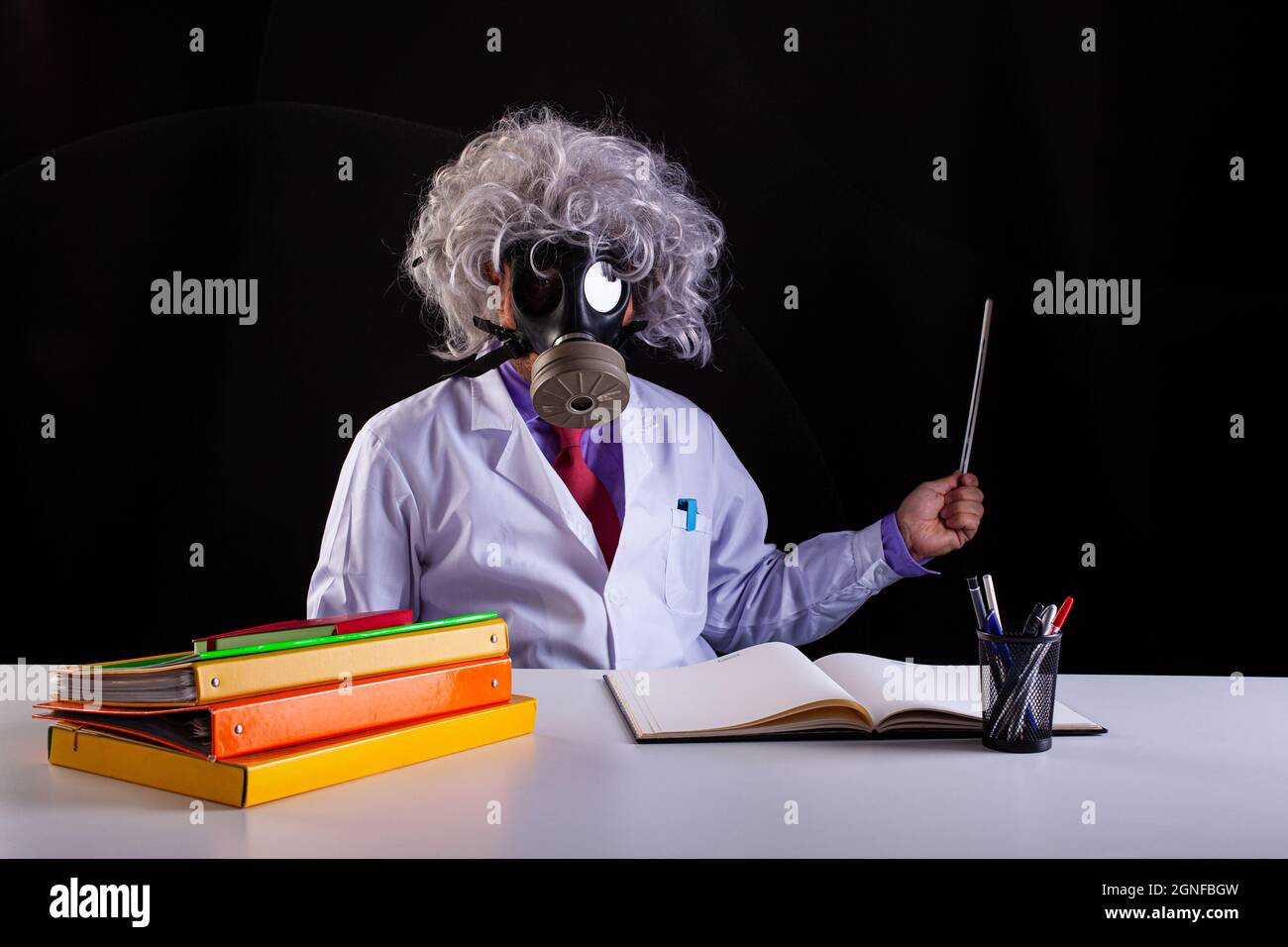 Pazzo insegnante di scienza in cappotto bianco con capelli unkempt seduto alla scrivania indossa una maschera a gas Foto Stock