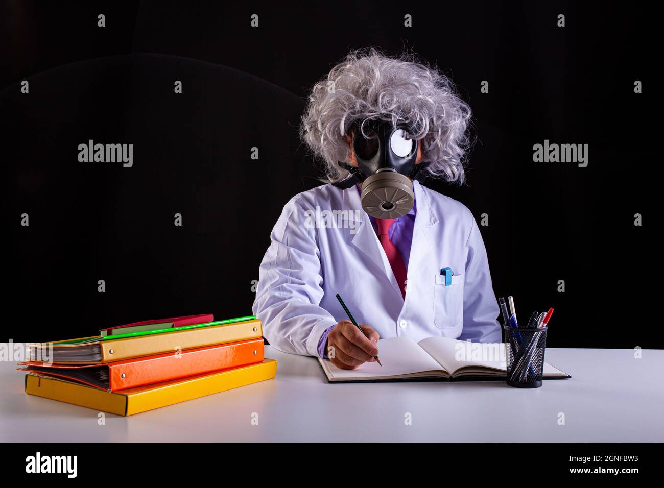 Pazzo insegnante di scienza in cappotto bianco con capelli unkempt seduto alla scrivania indossa una maschera a gas Foto Stock