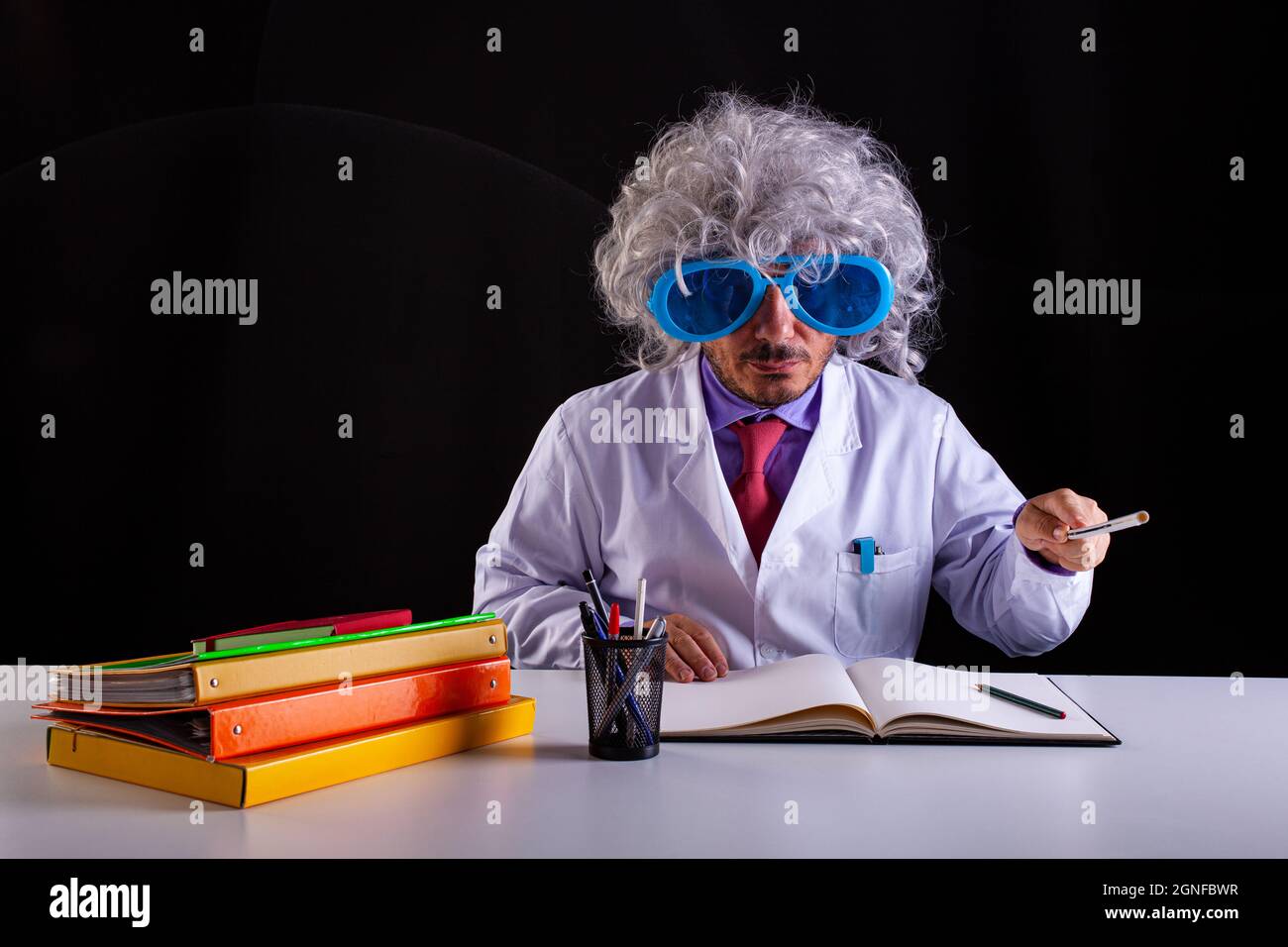 Insegnante di scienza pazzesca in cappotto bianco con capelli unkempt in divertenti occhiali da vista seduti alla scrivania Foto Stock