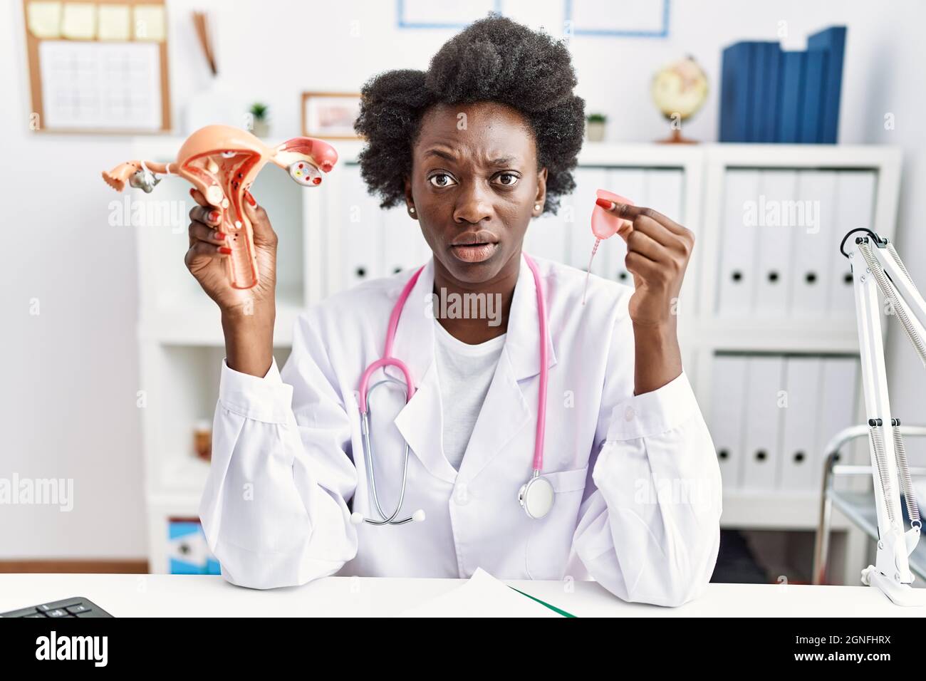 Medico africano donna che tiene anatomico organo genitale femminile e tazza mestruale Clueless e confuso espressione. Concetto di dubbio. Foto Stock