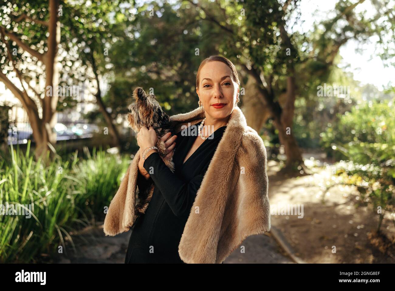 Spensierata donna elegante guardando la macchina fotografica mentre tiene il suo cucciolo all'aperto. Donna di fascia alta di età media in piedi da sola in un parco durante il giorno. FEMA Foto Stock