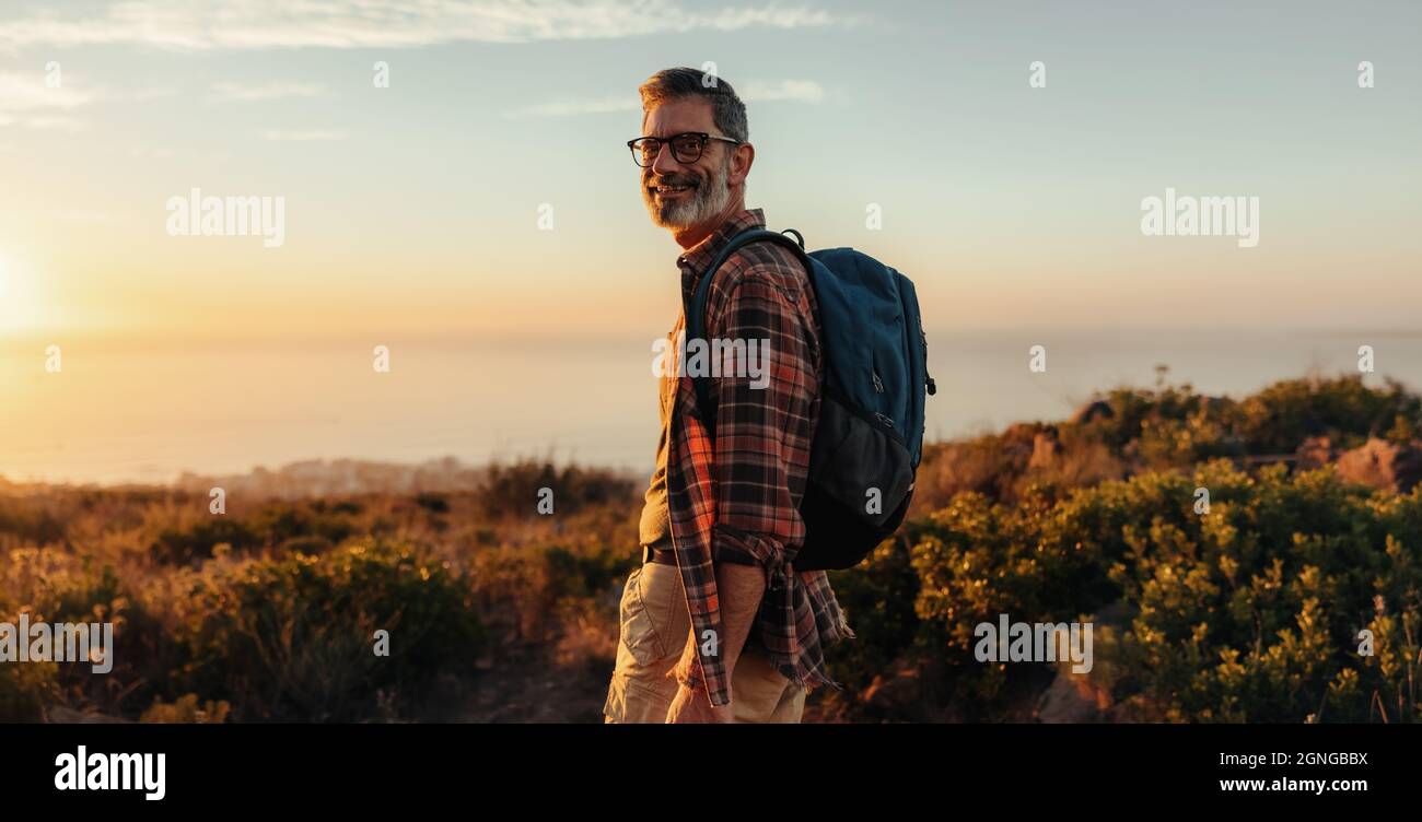 Happy escursionista maschile sorridendo alla macchina fotografica mentre si è in piedi da solo. Uomo maturo e allegro che porta uno zaino e si trova in cima a una collina. Zaino avventuroso Foto Stock