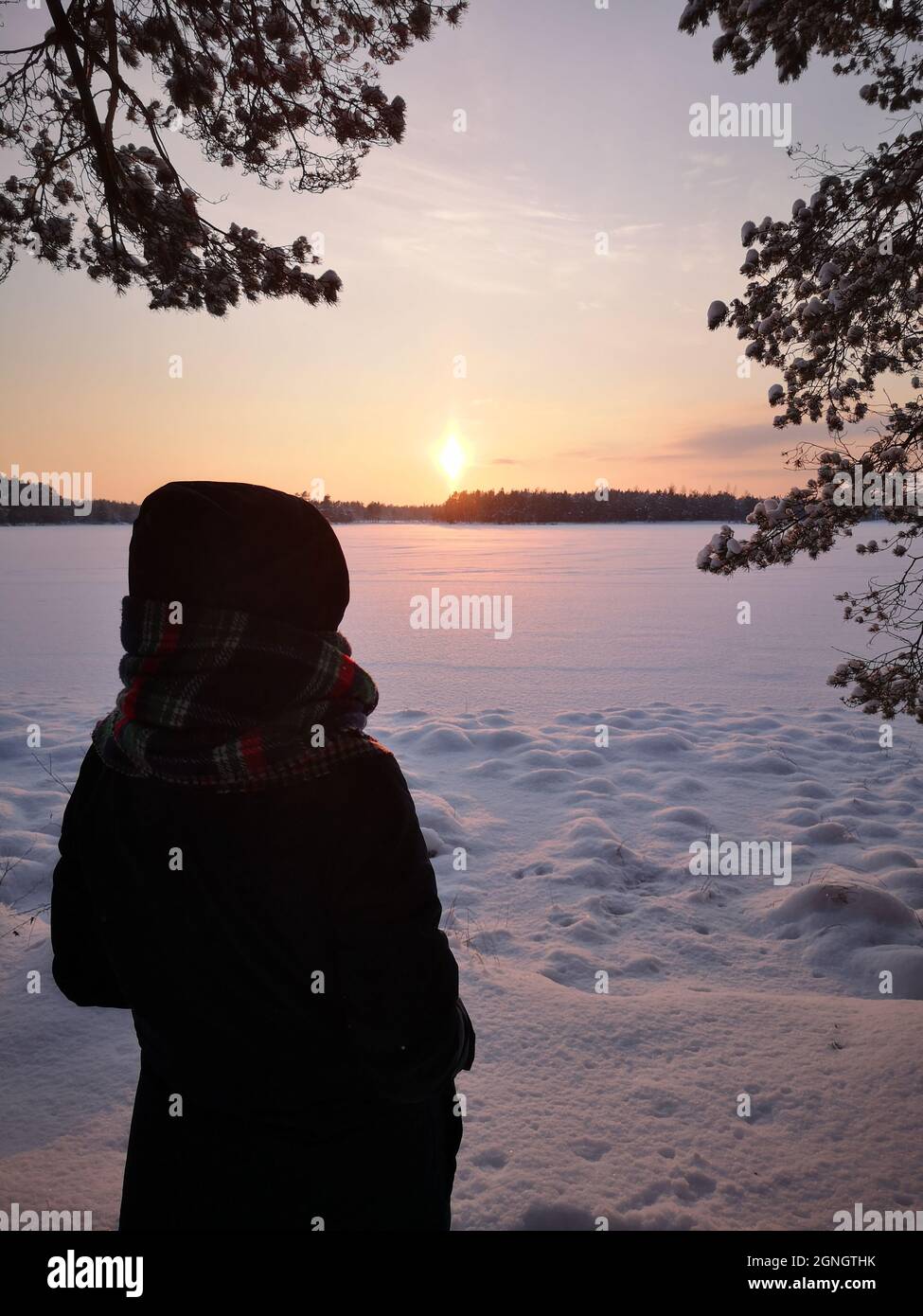 Donne in una giacca puffer e una sciarpa calda che guarda il fenomeno delle colonne di luce durante il tramonto su un lago ghiacciato in una serata invernale in Lettonia Foto Stock