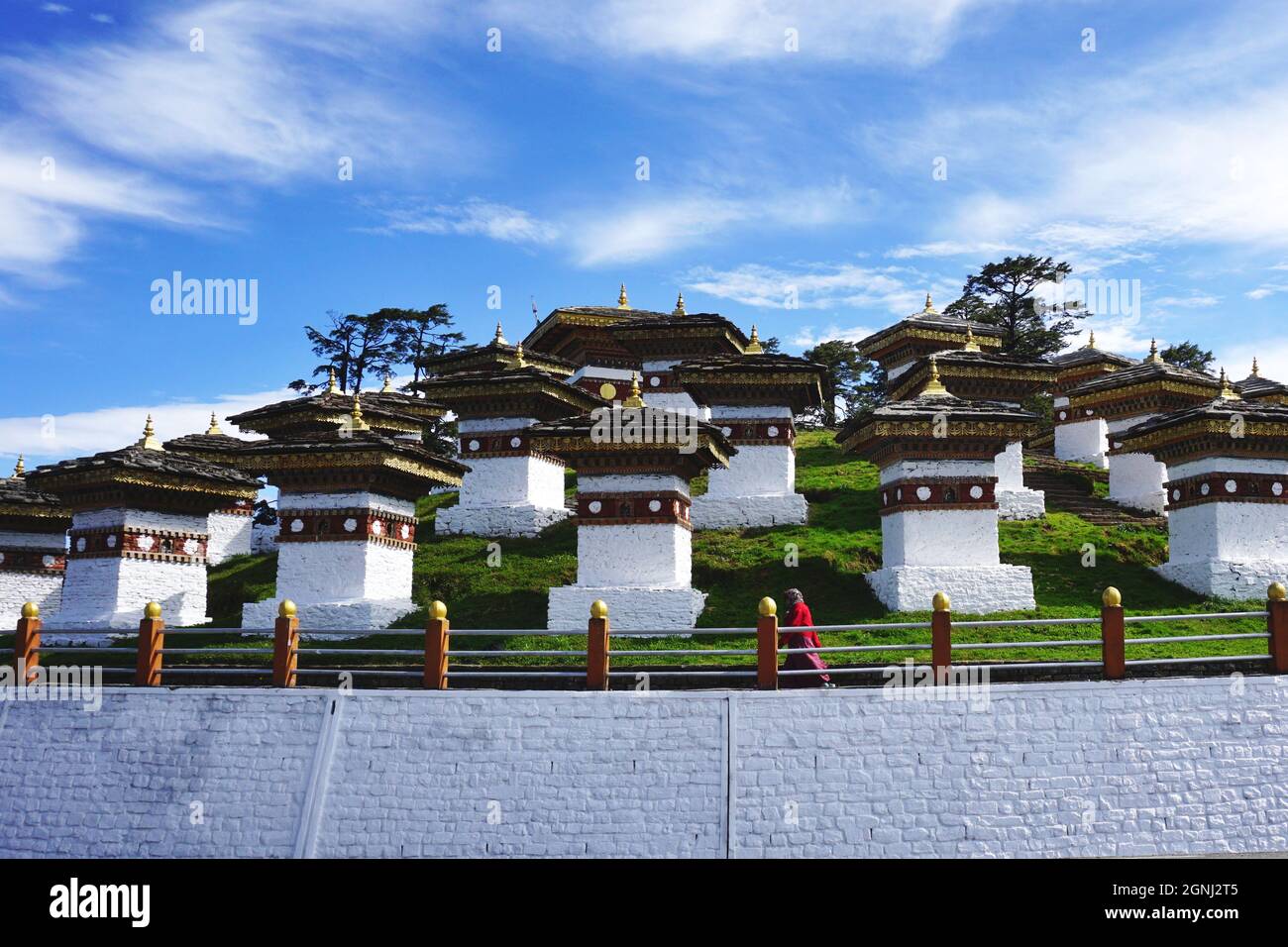 Una donna solista in rosso cammina davanti ad alcuni dei 108 Chortens Druk Wangyal identici che si trovano sul Passo Dochula di Bhutan sulla strada da Thimphu a Punakha. Foto Stock