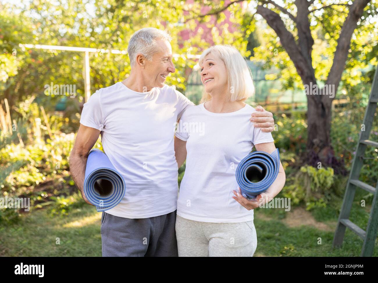 Sposi anziani attivi felici che tengono stuoie di yoga, esercitandosi insieme all'aperto nel loro giardino, spazio di copia Foto Stock