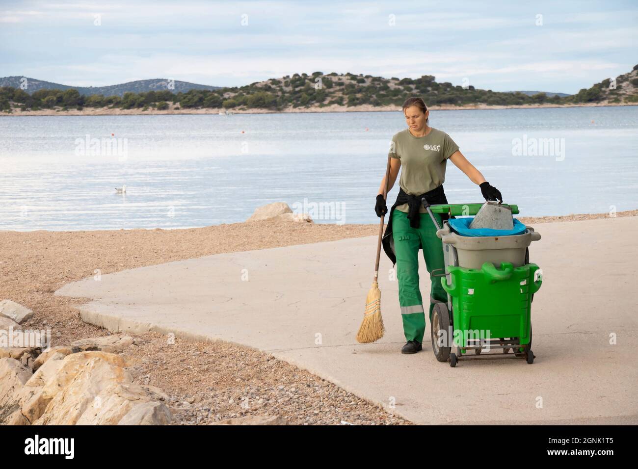 Vodice, Croazia - 28 agosto 2021: Giovane donna più pulita, che lavora per il servizio cittadino di Lec, al suo servizio di routine mattina pulizia della spiaggia Foto Stock