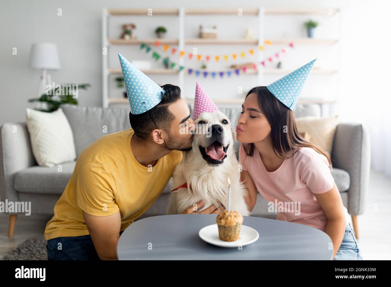 Giovane coppia interrazziale in cappelli di partito che celebra il compleanno del loro cane, baciando il loro animale domestico a casa Foto Stock