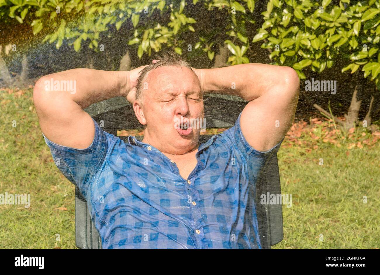 Ritratto di uomo maturo felice sotto spruzzi d'acqua in una giornata calda in giardino. Foto Stock