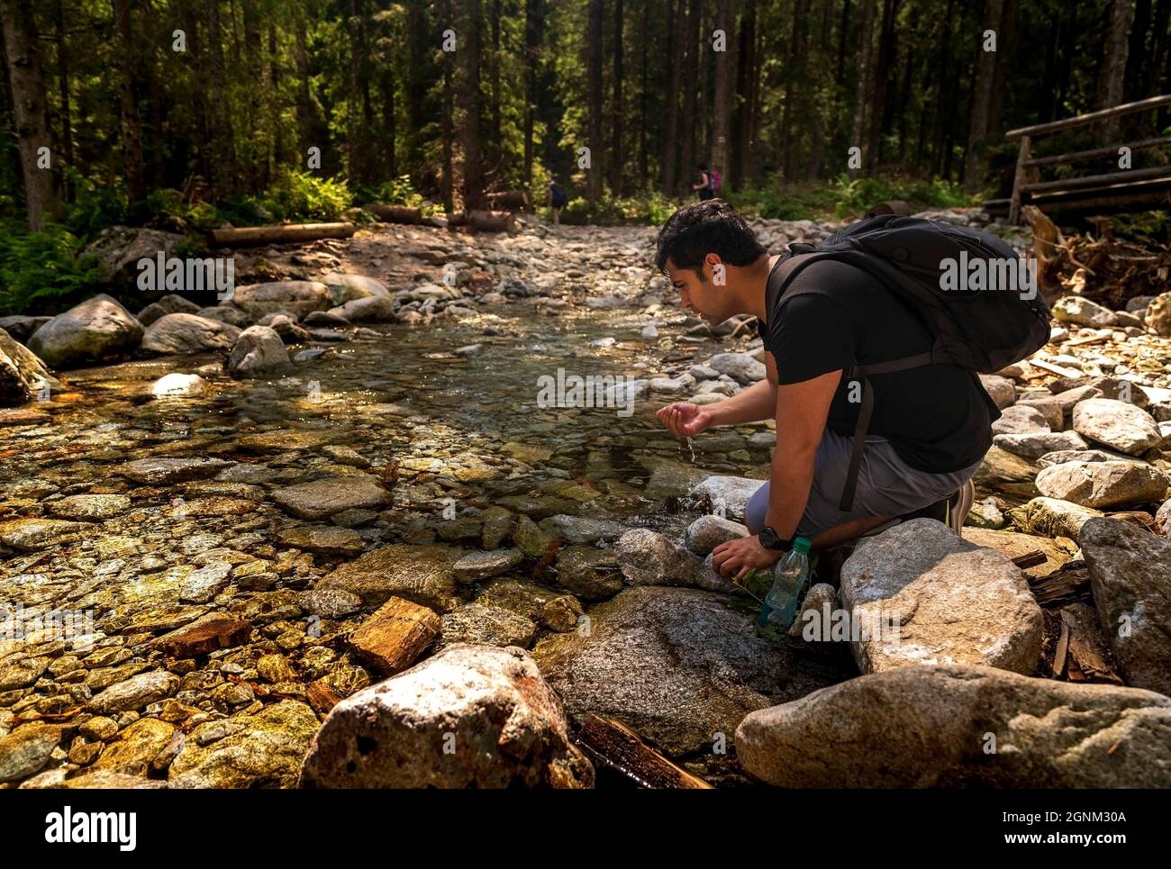 Uomo arabo indiano marrone con zaino seduto accanto al flusso d'acqua pulita o acqua potabile del fiume da esso, facendo una pausa durante il trekking o un'escursione nella foresta Foto Stock