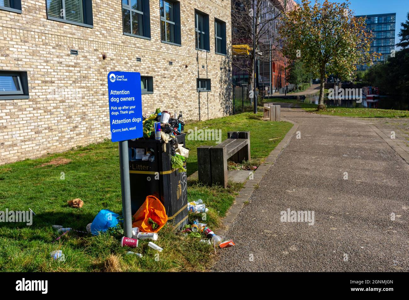 Segno che chiede ai proprietari del cane di prendere dopo i loro cani, accanto ad un contenitore di rifiuti imballato e traboccante. Da Ashton Canal, Ancoats, Manchester, Inghilterra, Regno Unito Foto Stock