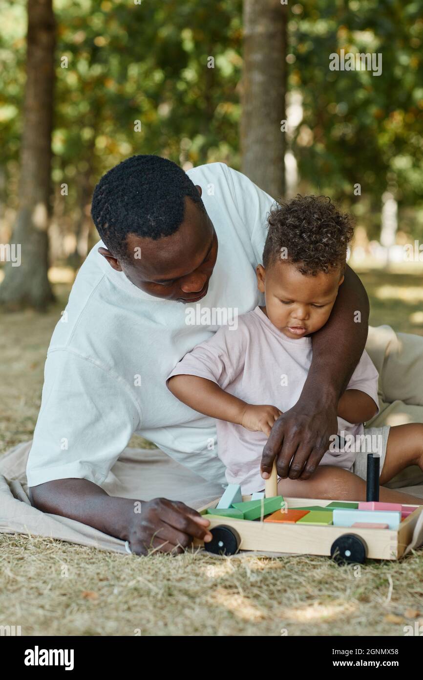 Ritratto verticale del giovane padre afroamericano che gioca con il figlio carino nel parco mentre si siede sull'erba Foto Stock