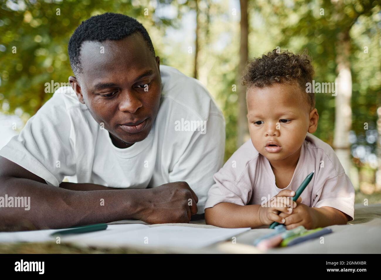 Ritratto della vista frontale di giovane padre afroamericano che gioca con un figlio carino nel parco mentre si posa sull'erba Foto Stock