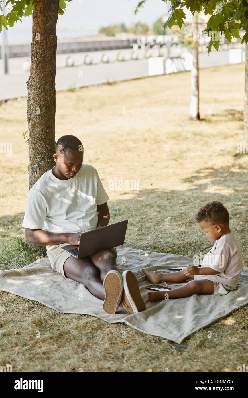 Ritratto verticale a tutta lunghezza di padre afro-americano usando il laptop mentre gioca con il figlio carino nel parco, spazio di copia Foto Stock