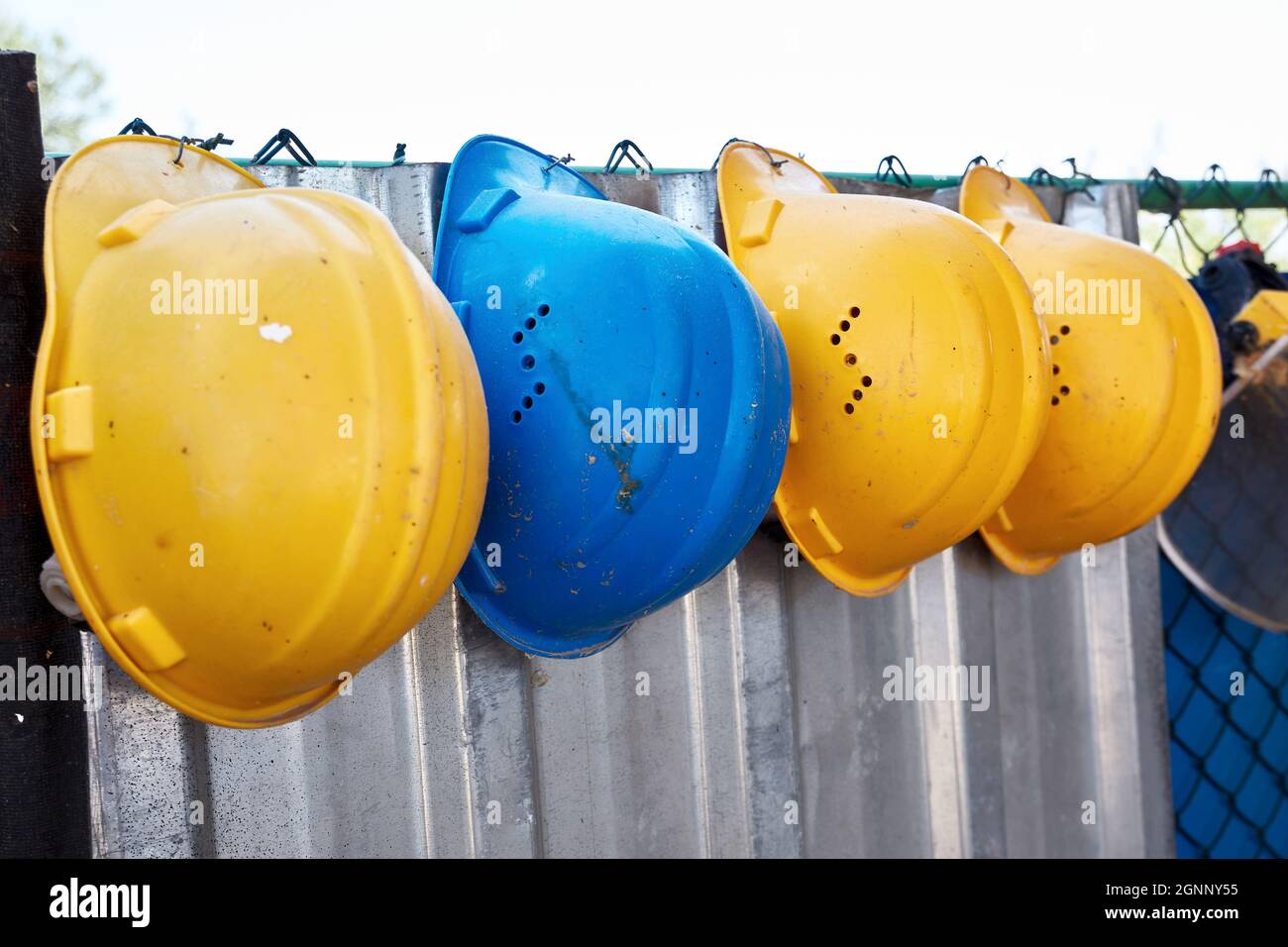 Cappello di sicurezza blu con cappelli di sicurezza gialli appesi su una recinzione in fila presso il cantiere. Sicurezza sul lavoro. Foto Stock