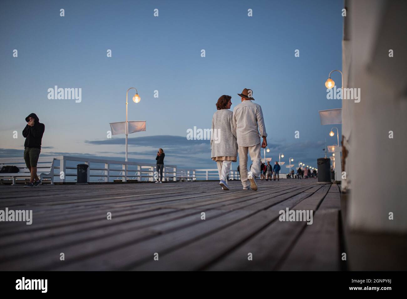 Vista posteriore ad angolo basso della coppia anziana felice che cammina all'aperto sul molo dal mare al crepuscolo, tenendo le mani. Foto Stock