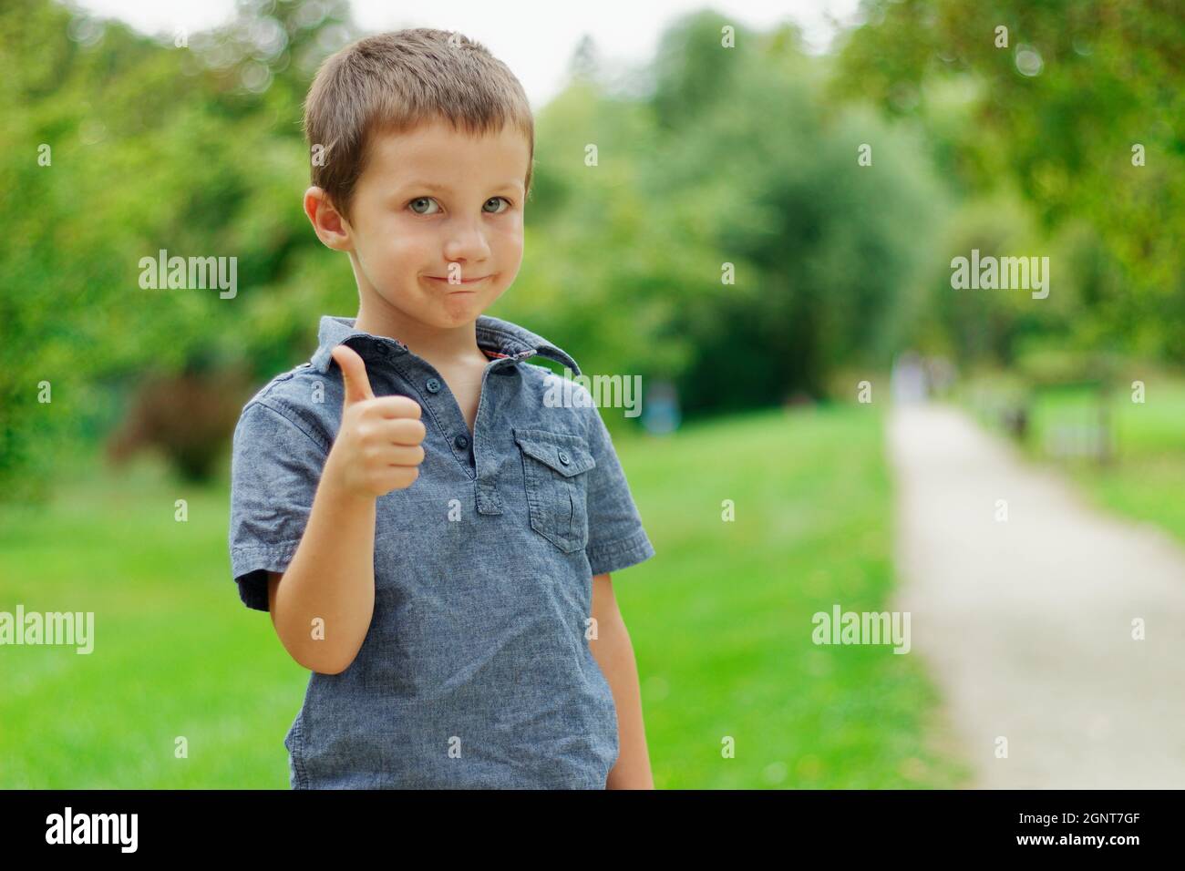 Ritratto di un ragazzo carino in un parco estivo verde. Il ragazzo biondo con gli occhi blu mostra il pollice in su Foto Stock