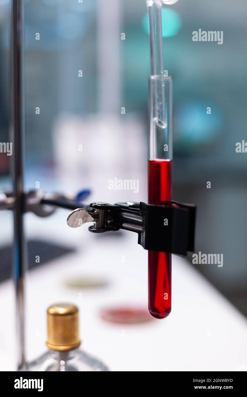 Primo piano della provetta con sangue sul tubo di gocciolamento nel laboratorio scientifico. Matraccio per vetreria chimica con liquido rosso e soluzione su banco da laboratorio. Ambiente di lavoro biochimico con strumenti Foto Stock