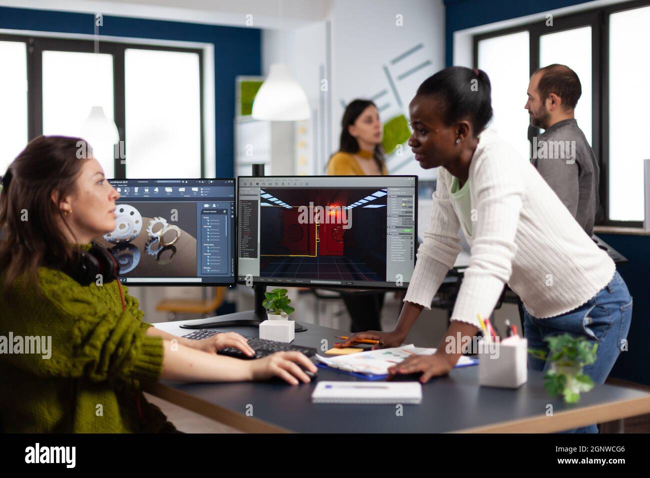 Designer multietnico che guarda computer con doppio display che lavorano insieme al progetto di gioco in ufficio aziendale. Lavoratori dei videogamer che sviluppano nuove applicazioni di gioco su pc con tecnologia moderna Foto Stock