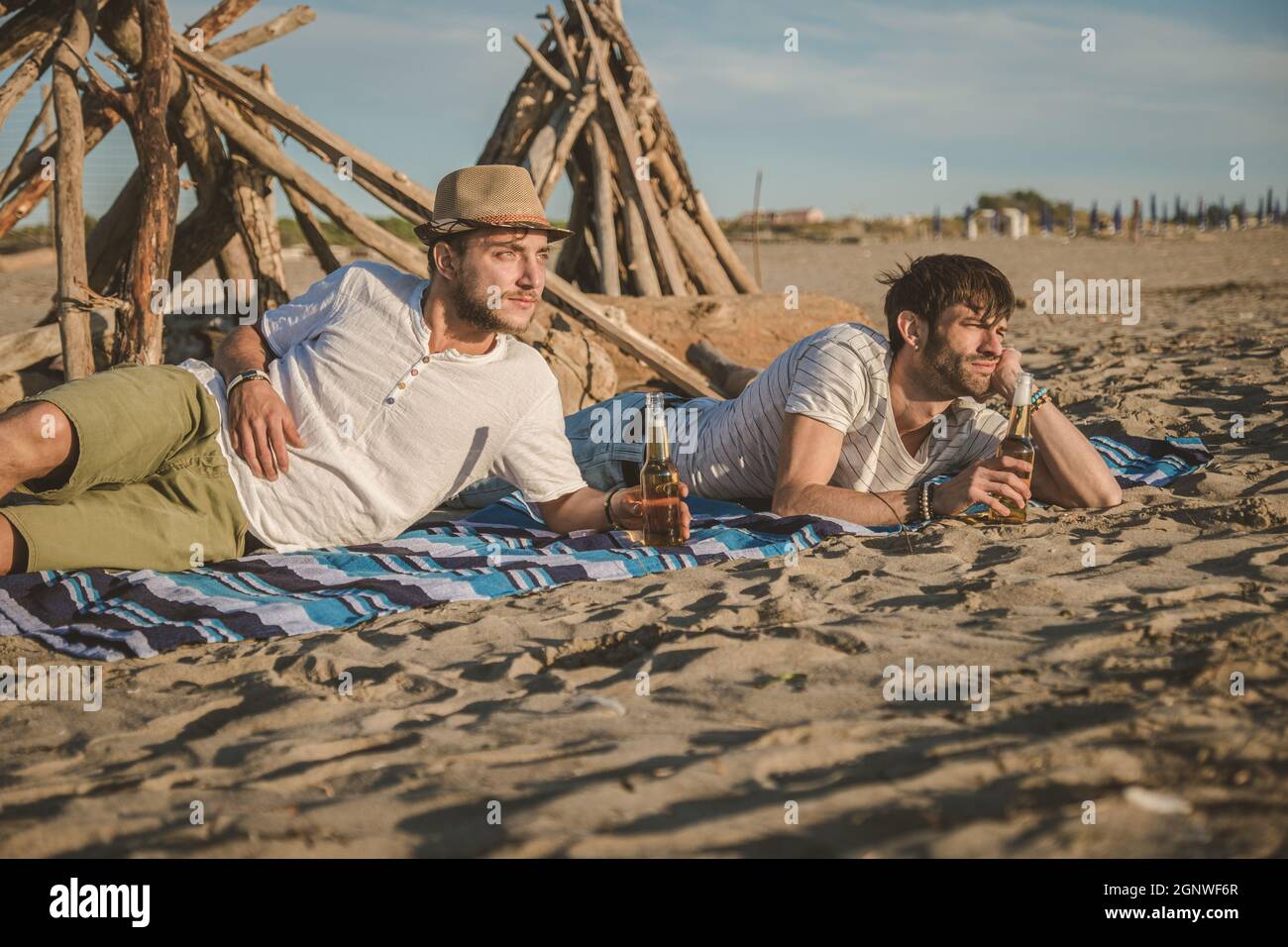 Due giovani amici trascorrono il tempo insieme seduti sulla spiaggia, bevendo birra e tostando in vacanza al tramonto estivo al crepuscolo. Concetto di amicizia Foto Stock
