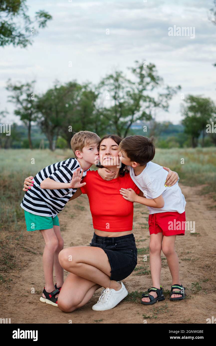 Autentico ritratto di famiglia a tutta lunghezza. Mamma casual giovane con due figli che abbraccia e bacia. Madre e bambino che camminano in campagna. Vite quotidiane. Attività quotidiane. Foto Stock