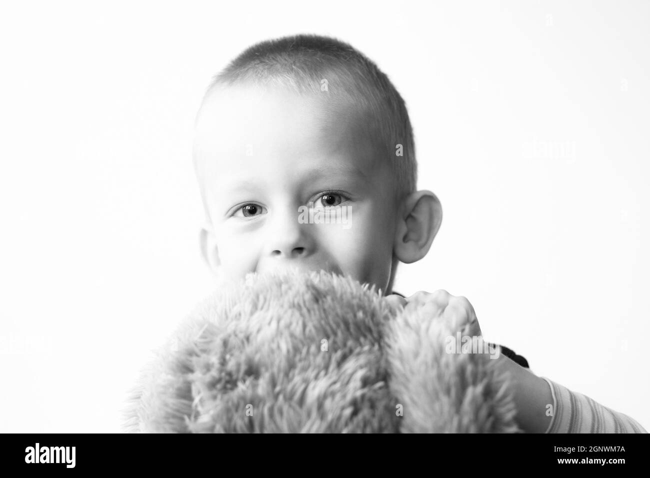 ragazzo di tre anni in posa con i suoi orsacchiotti che ne abbraccia uno e guarda la macchina fotografica e sorride contro il muro bianco, immagine in bianco e nero Foto Stock