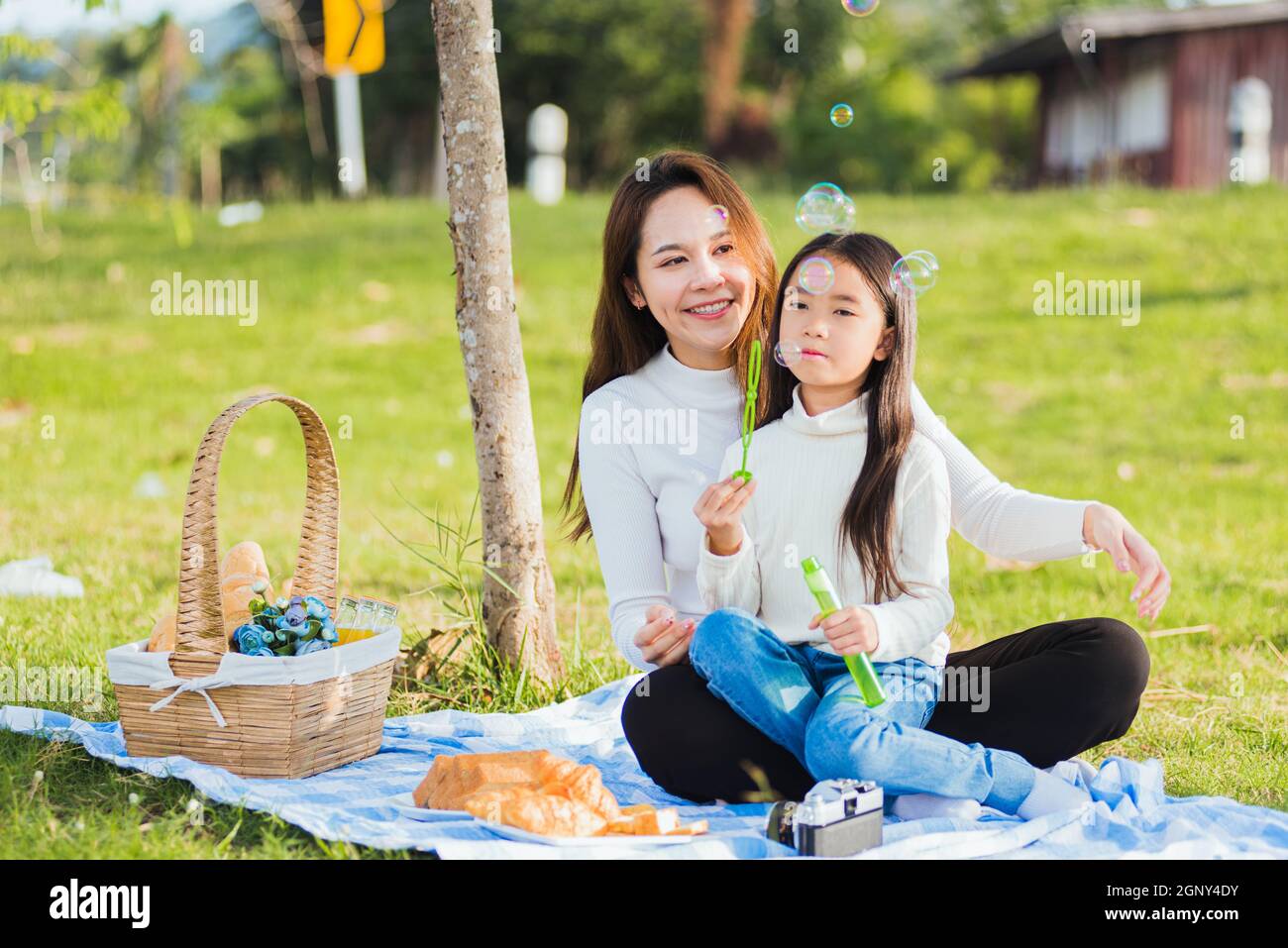 Buona madre asiatica e bambina bambina che si divertono e godendo all'aperto insieme seduto sul sapone che soffia l'erba bolle durante un picnic i Foto Stock