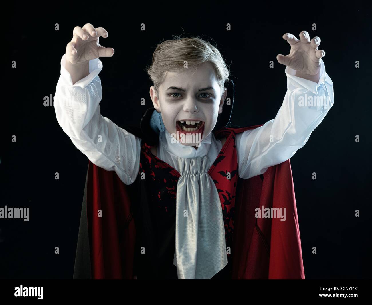 Ritratto di ragazzo indossando vampiro di Halloween trucco e costume mantello ha beffato i denti, isolato su sfondo nero. Foto Stock