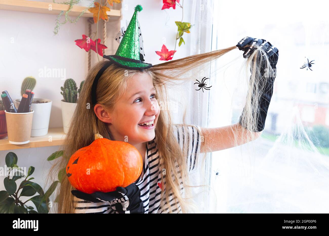 Ragazza caucasica allegra in un cappello di strega seduto su una finestra decorata per Halloween, tenendo una zucca, ragni sedersi in una rete di capelli. Preparazione in corso Foto Stock