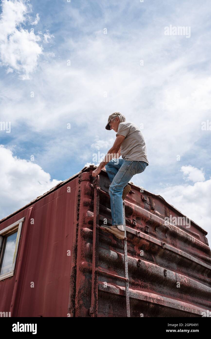 Guarda un uomo anziano vestito con jeans blu e t-shirt che sale su una scala di metallo sul retro di un vagone rosso, sostenuto da cielo blu e nuvole. Foto Stock