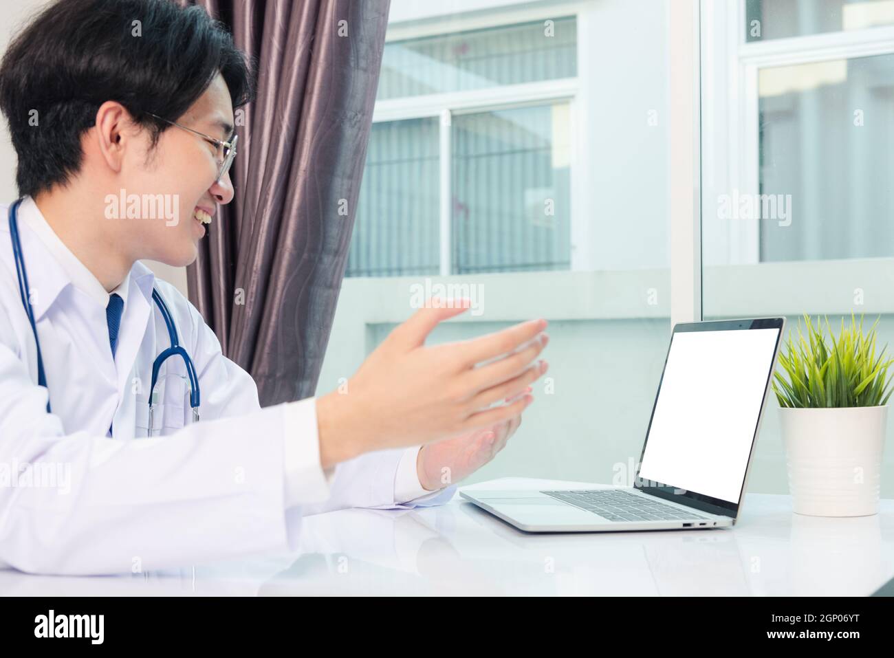 Giovane e bello medico asiatico che indossa un abito medico e stetoscopio videoconferenza chiamata o facetime sollevare la mano per spiegare i sintomi che smili Foto Stock