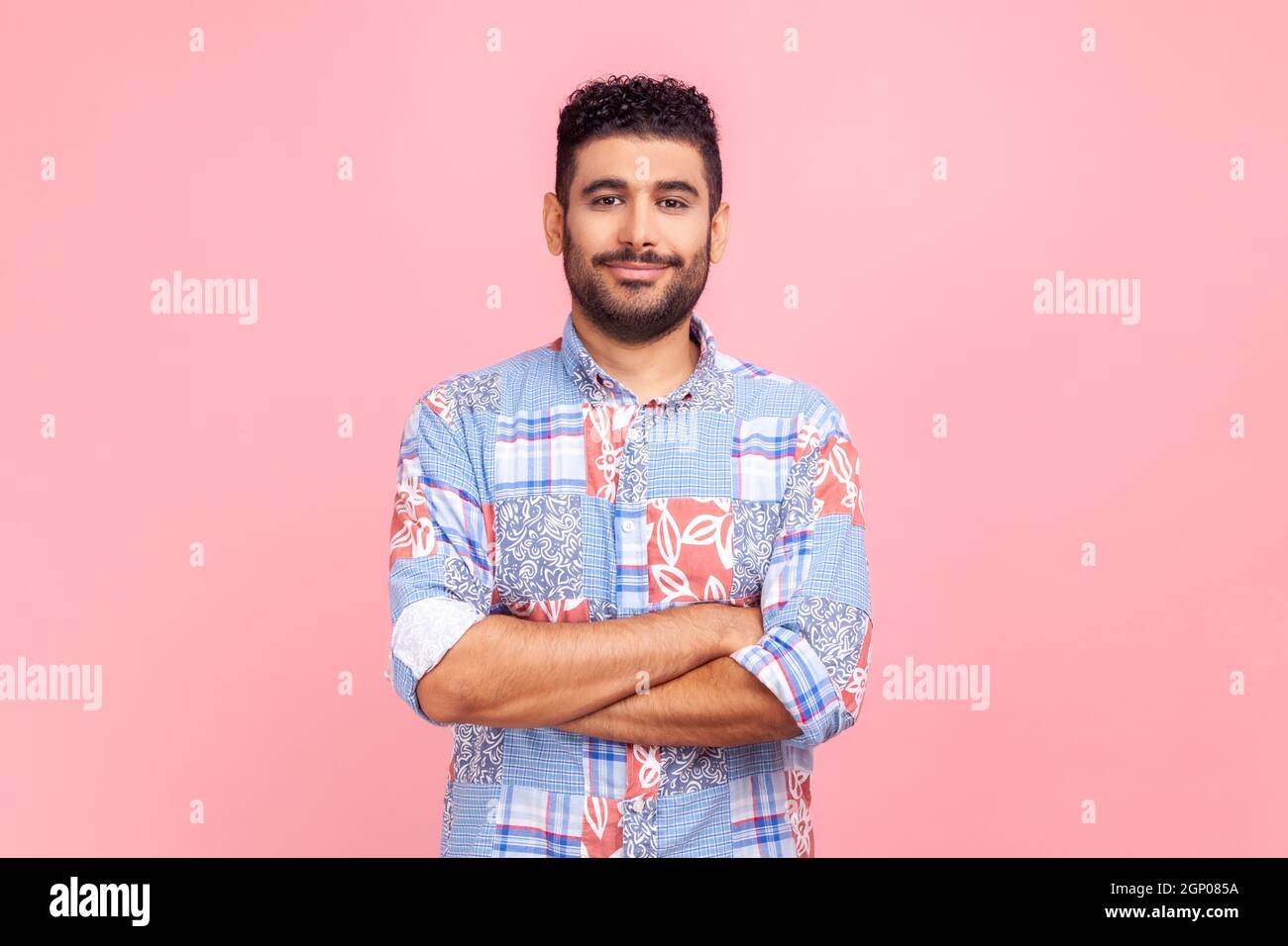 Uomo sorridente con camicia blu casual in piedi con le mani ripiegate e guardando la fotocamera con espressione facciale sicura. Studio interno girato isolato su sfondo rosa. Foto Stock