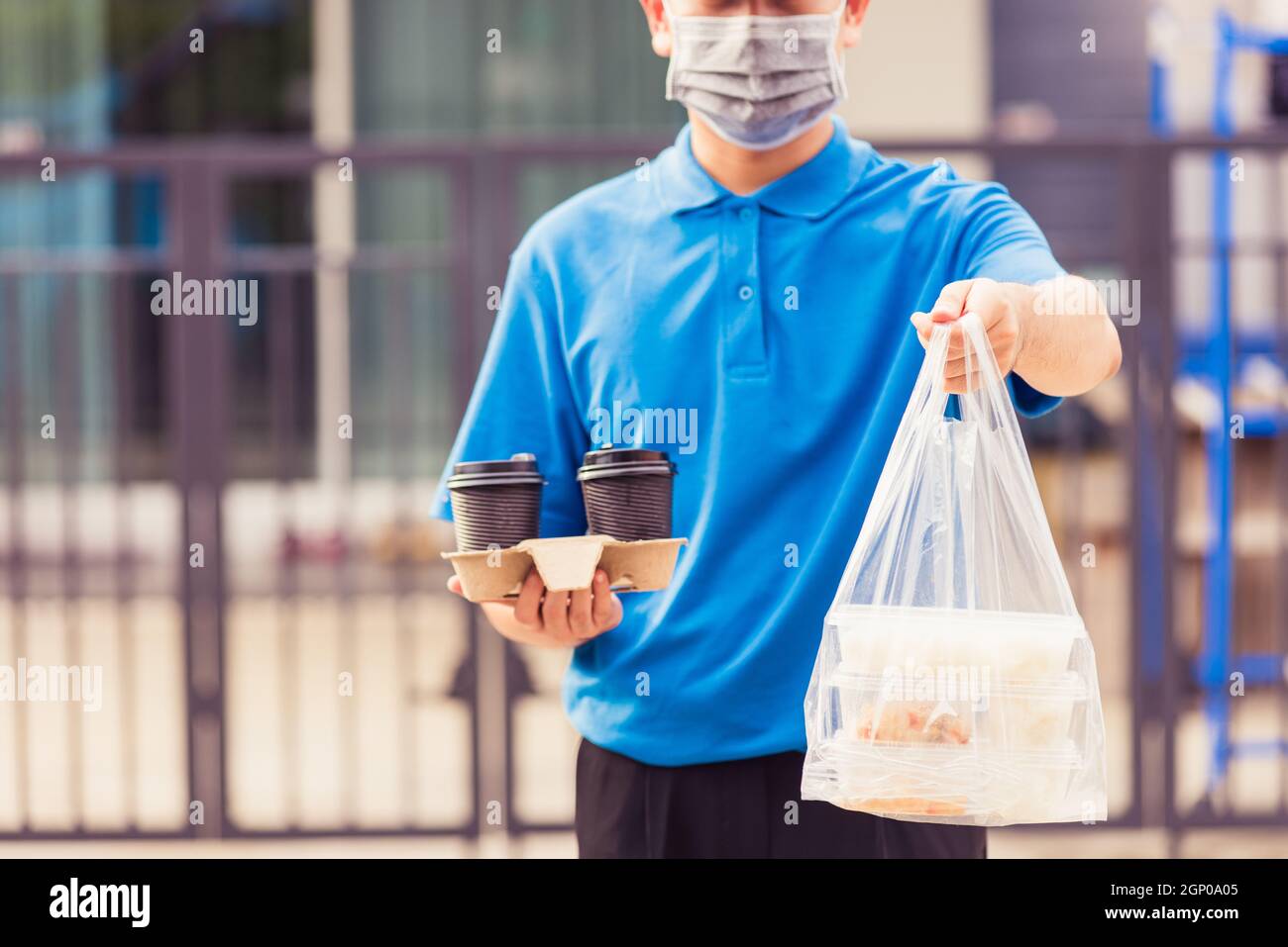 Giovane uomo asiatico di consegna in uniforme blu maschera di indossare faccia servizio di drogheria che dà riso scatole di plastica sacchetti di cibo e caffè in casa davanti sotto Foto Stock