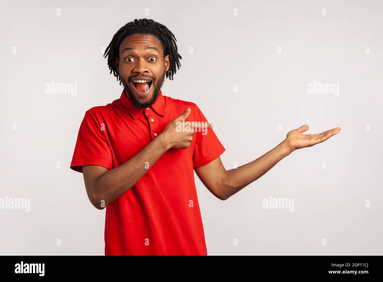 Bell'uomo con dreadlock che indossa una T-shirt rossa, che presenta uno spazio per la copia sul palmo e che guarda sorpreso, mostrando spazio vuoto per il testo promozionale. Studio interno girato isolato su sfondo grigio. Foto Stock