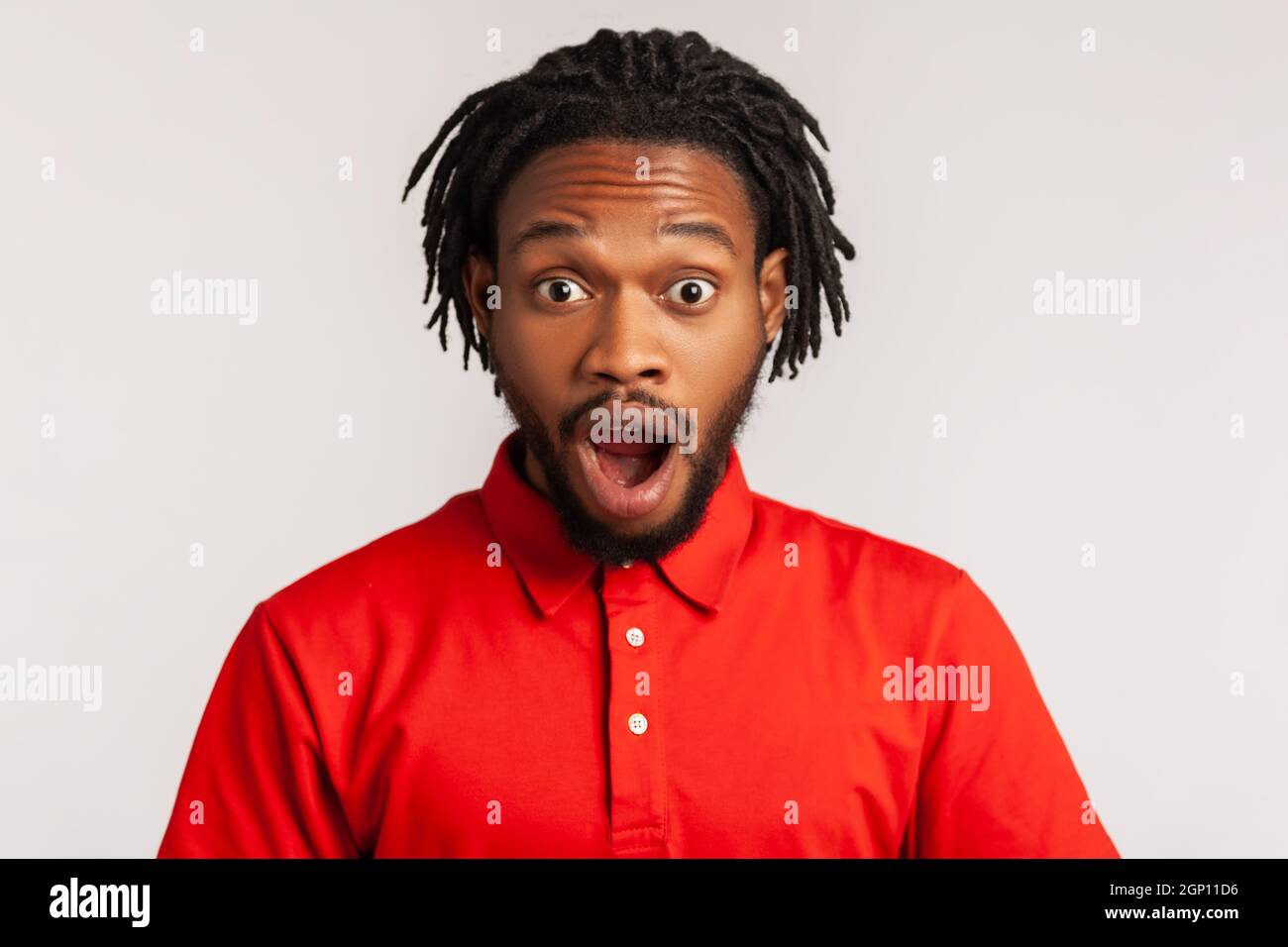 L'uomo stupito con i dreadlock che indossano una T-shirt rossa casual, in piedi con la bocca aperta di sorpresa, ha shockato l'espressione, sente le notizie incredibili. Studio interno girato isolato su sfondo grigio. Foto Stock