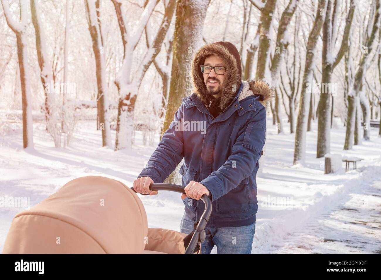 Il padre felice cammina attraverso il parco in inverno con una carrozza per bambini. Camminando con il bambino in inverno la mattina. Nevicate, concetto di Blizzard. Foto Stock