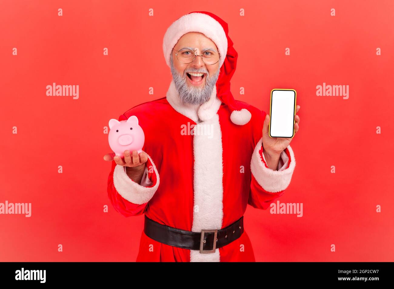 Uomo anziano sorridente con barba grigia in costume babbo natale in piedi tenendo smartphone con schermo bianco e bianco e Piggy banca, cashback, portafoglio online. Studio interno girato isolato su sfondo rosso. Foto Stock