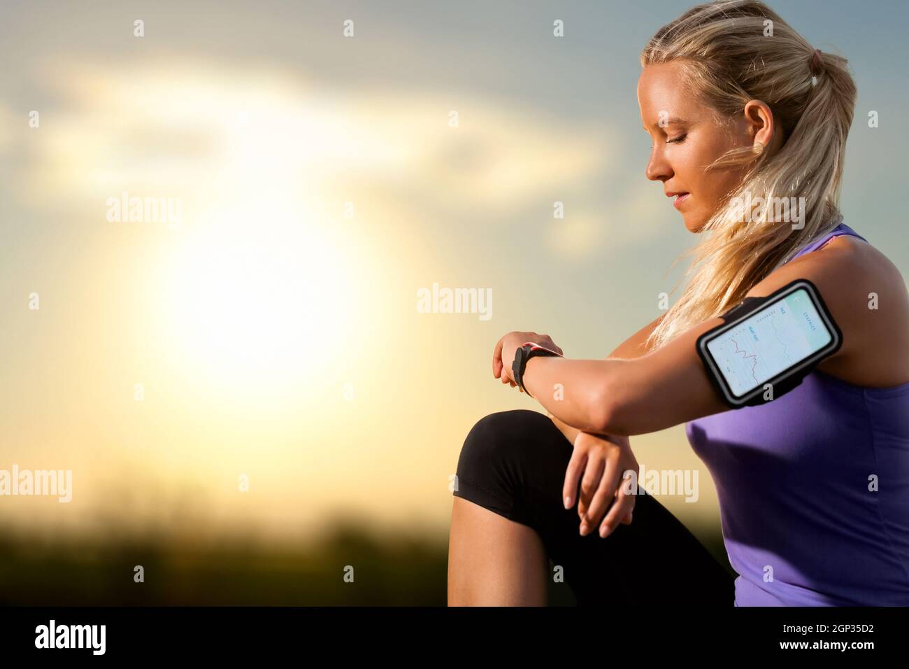 Ritratto del giovane atleta che controlla l'allenamento su un orologio intelligente al tramonto. Donna che indossa una fascia da braccio con orologio intelligente e grafica che mostra i risultati dell'allenamento. Foto Stock