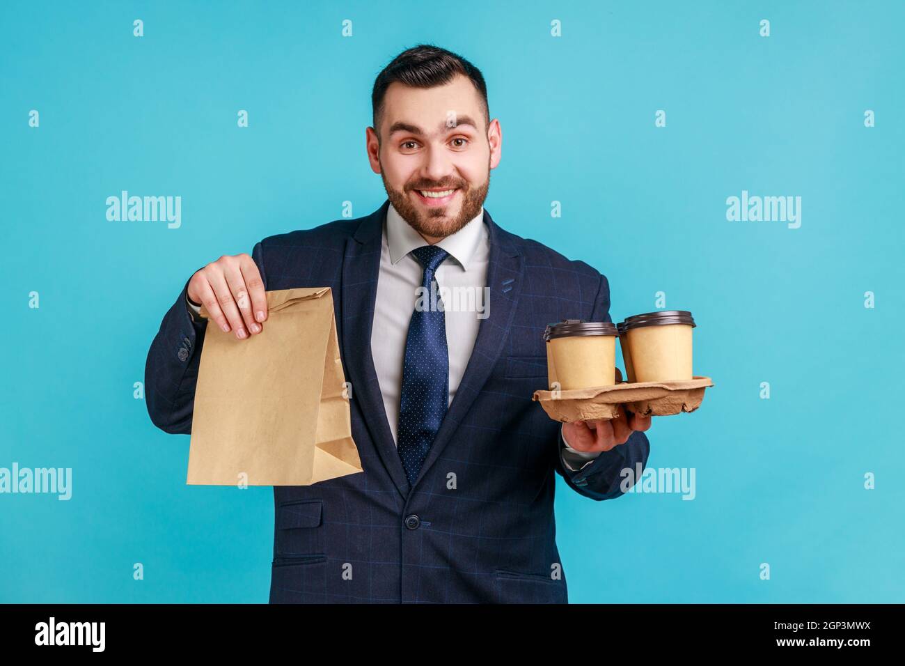 Felice uomo sorridente con portico vestito ufficiale che tiene carta pacchetto e portare via il caffè in mani, consegna rapida, colazione. Studio interno girato isolato su sfondo blu. Foto Stock