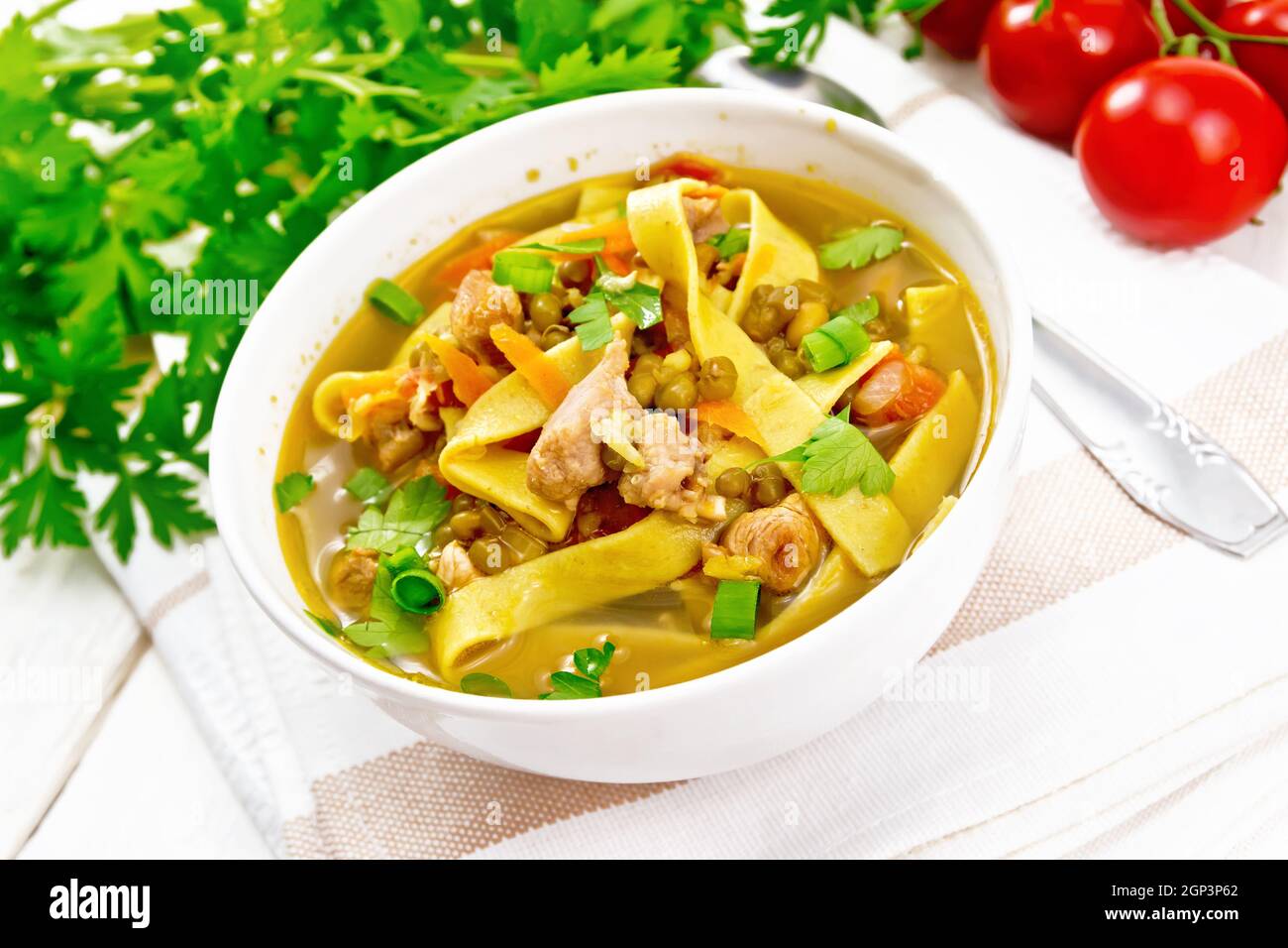 Zuppa con carne, pomodori, verdure, lenticchie di fagioli e tagliatelle in una ciotola su un tovagliolo, prezzemolo e cucchiaio sullo sfondo di una tavola di legno chiaro Foto Stock