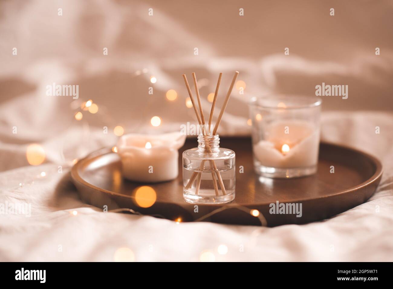 Profumo domestico in bottiglia di vetro con i bastoni di legno, candele di bruciatura profumate sul vassoio in camera da letto vicino su bianco. Aromaterapia atmosfera accogliente stile di vita. Wi Foto Stock